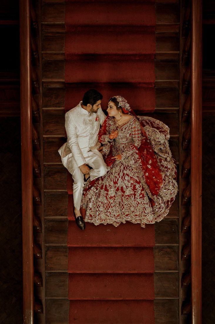 Overhead shot of bride and groom seated together on a red-carpeted wooden staircase, her embellished red lehenga spread across the steps