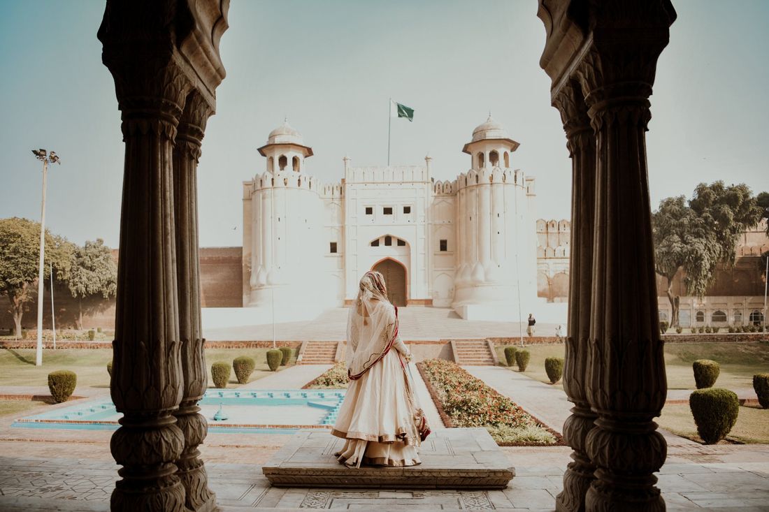 Bride viewed from behind looking across a garden toward the Alamgiri Gate of Lahore Fort, framed by carved wooden pillars
