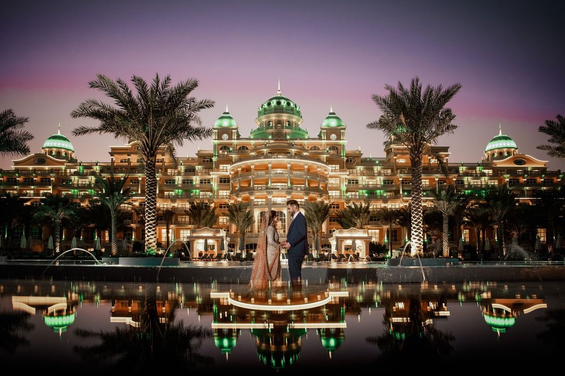 Bride and groom holding hands on a reflective pool platform at twilight, an illuminated palace-style resort with green domes behind them, destination wedding photography by Arfa & Usman