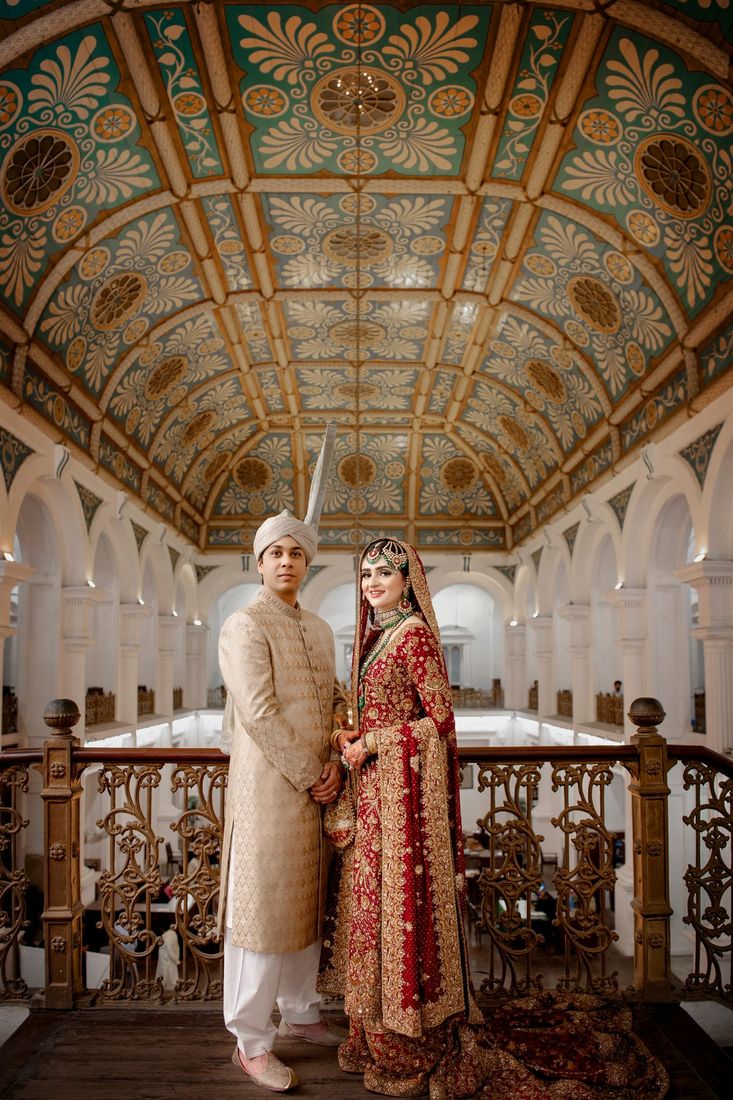 Bride and groom standing on a balcony beneath an ornately painted vaulted ceiling in a grand historic hall
