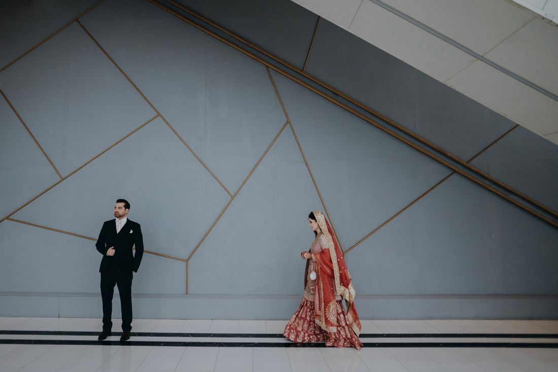 Bride walking past the groom against a geometric modern wall with gold inlay lines, an escalator visible above