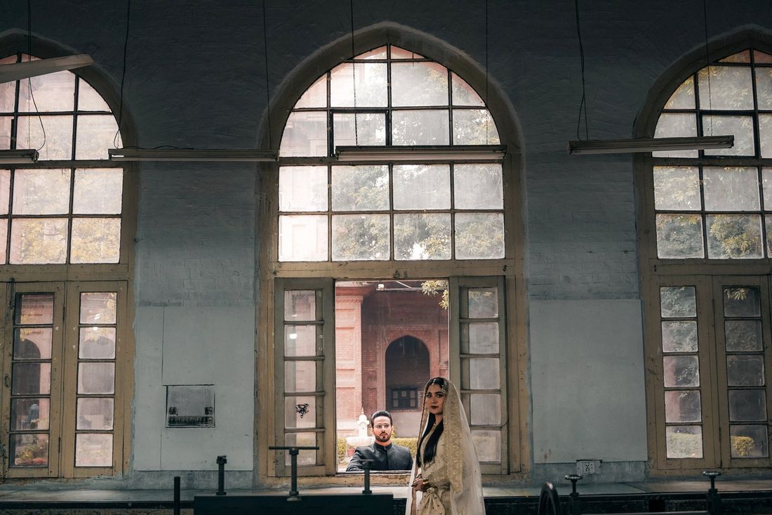 Bride walking through an arched doorway of a heritage building with tall industrial windows, groom visible in the background