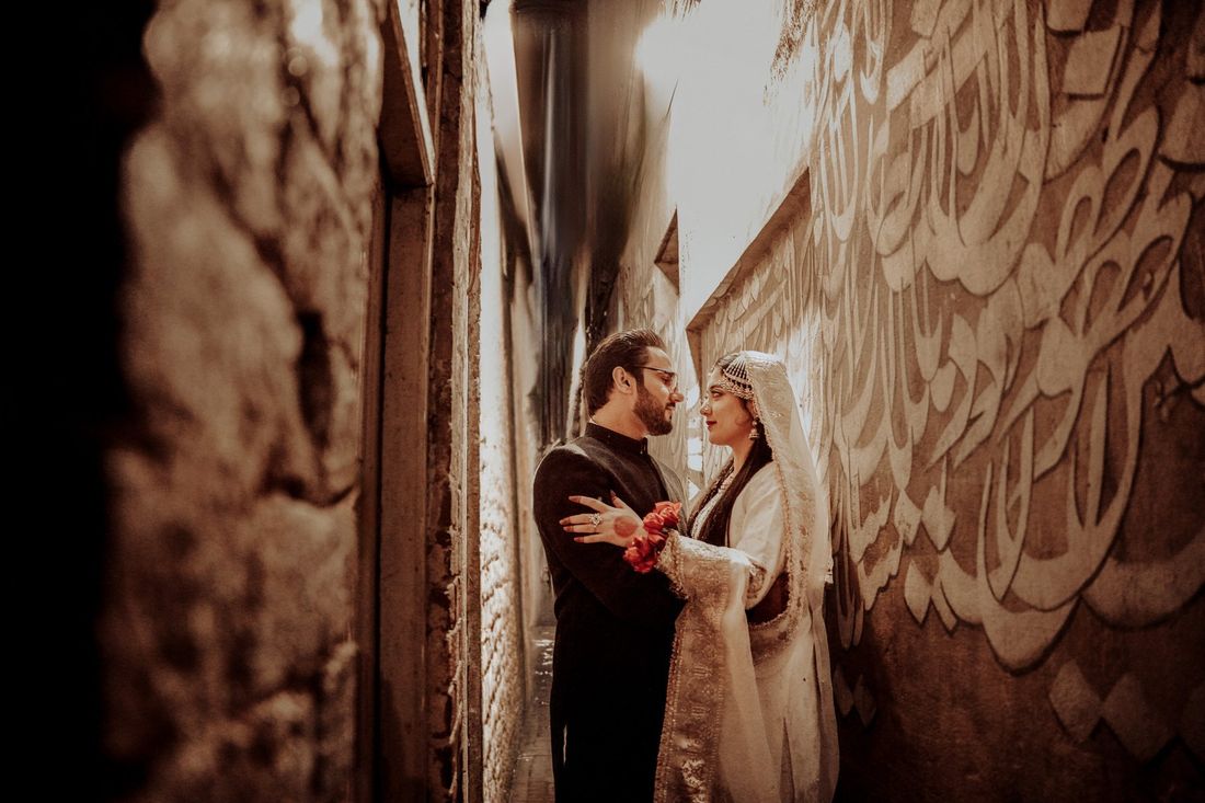 Bride and groom embracing in a narrow alley beside a wall painted with large Urdu calligraphy