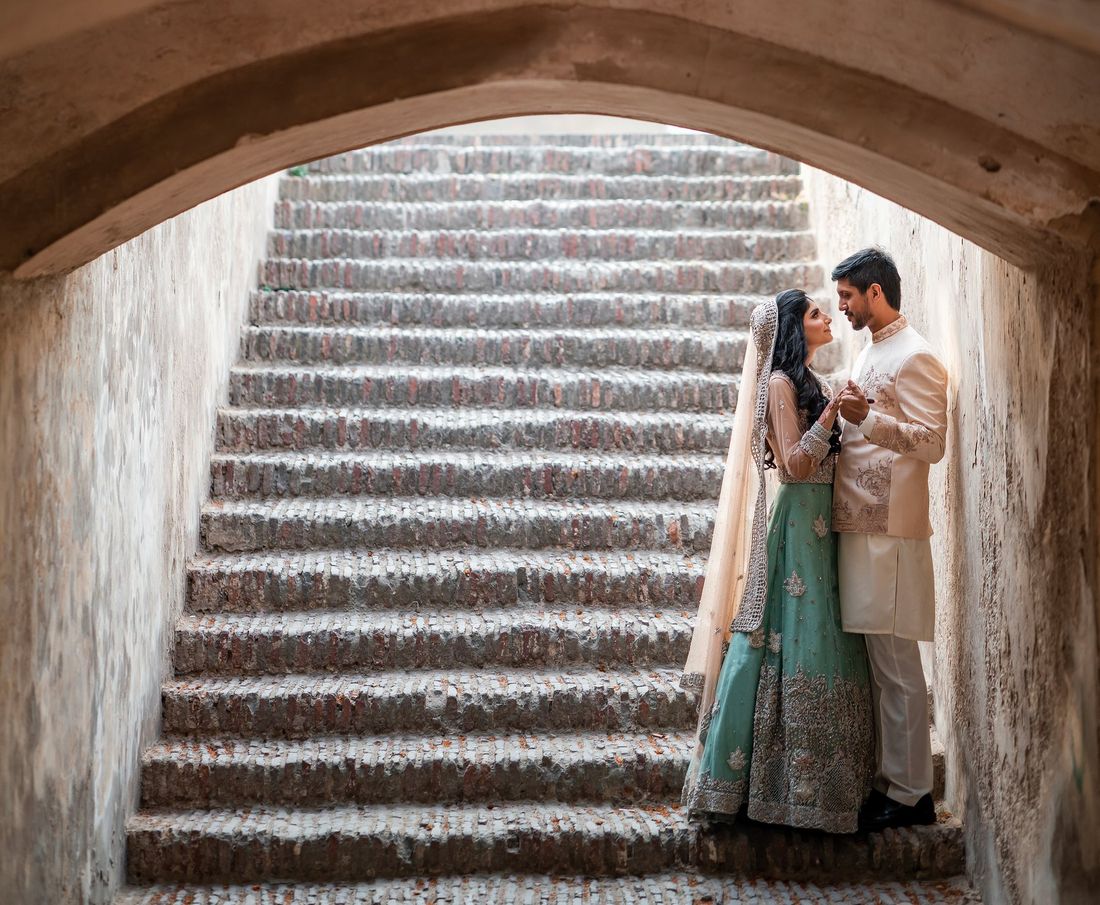 Bride in a sage green embellished lehenga and groom in a cream sherwani standing close at the base of a historic stone stairway