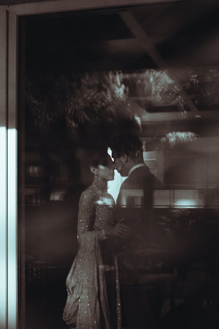 Bride and groom seen through a reflective glass window at night, their embrace softened by overlapping reflections