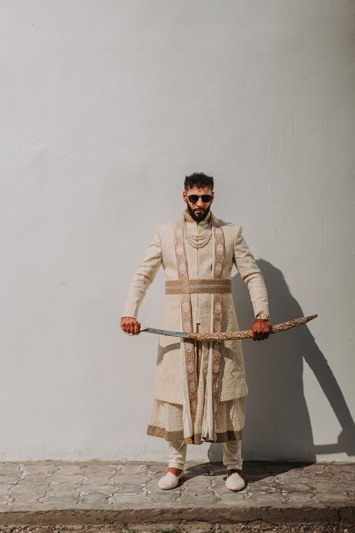 Groom in an ivory embellished sherwani holding a ceremonial sword, standing confidently against a plain white wall