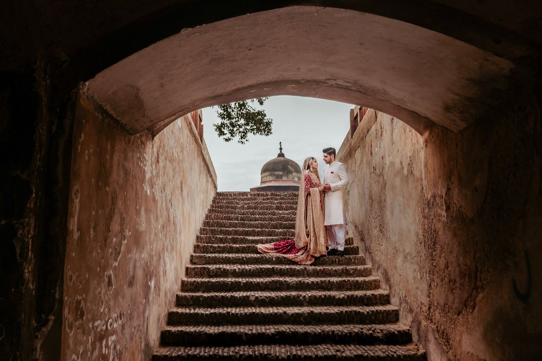 Bride and groom standing atop a historic stone stairway, viewed from below through an arched tunnel with a distant dome visible