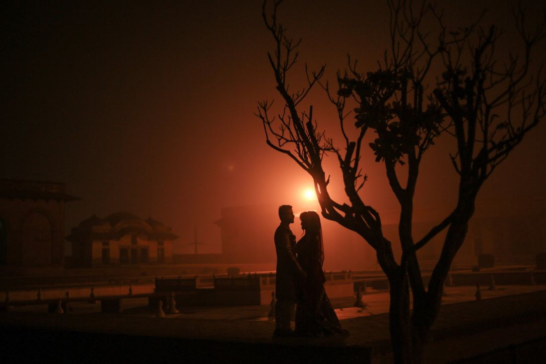 Silhouette of bride and groom embracing beneath a bare tree, glowing orange light behind them on a heritage rooftop at night