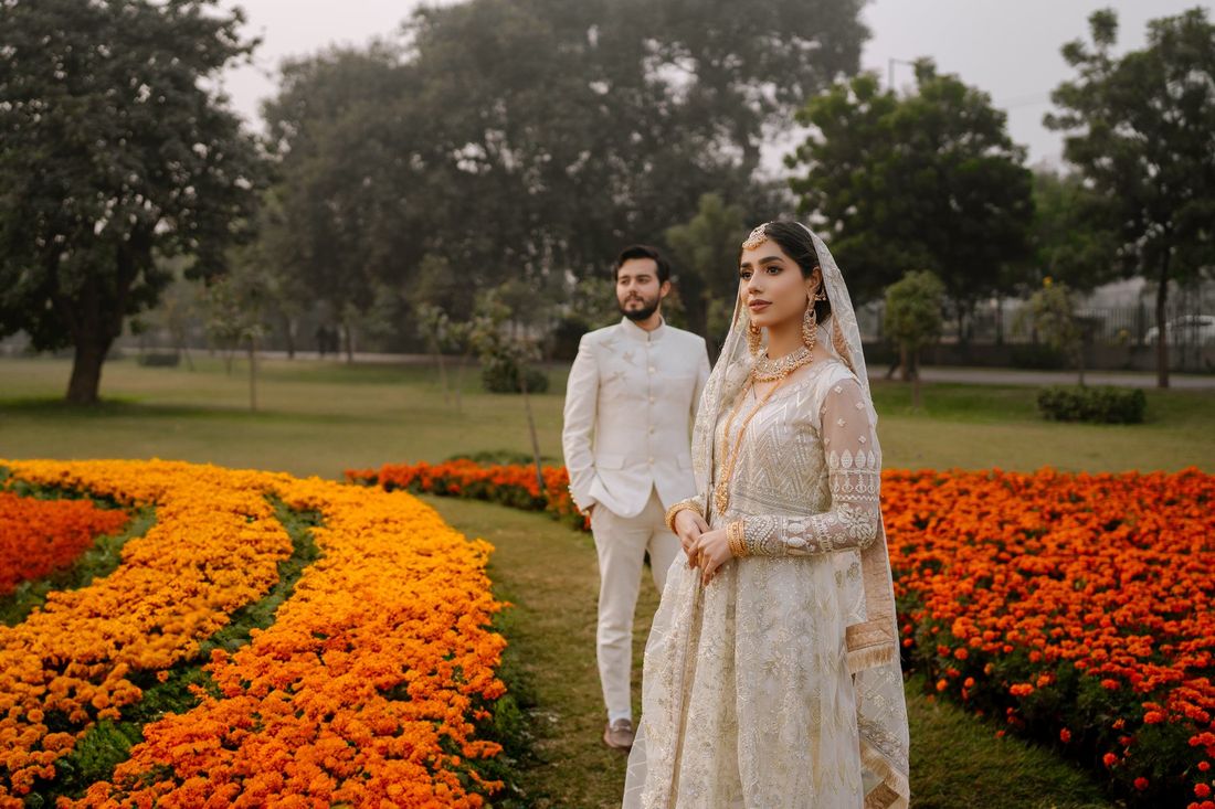 Bride standing before vivid orange and yellow marigold flower beds, groom walking behind her in the garden