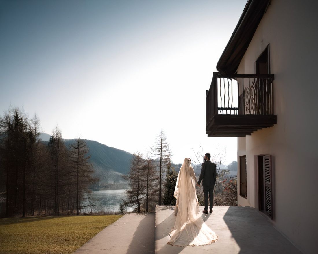 Bride and groom walking hand in hand on a terrace overlooking a mountain lake, viewed from behind