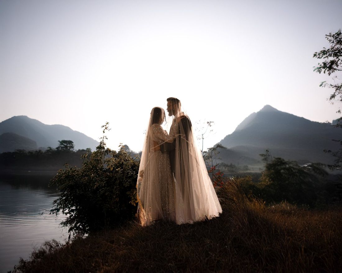 Bride's sheer veil draped over both her and the groom as they embrace beside a lake at golden hour, layered mountains in the background