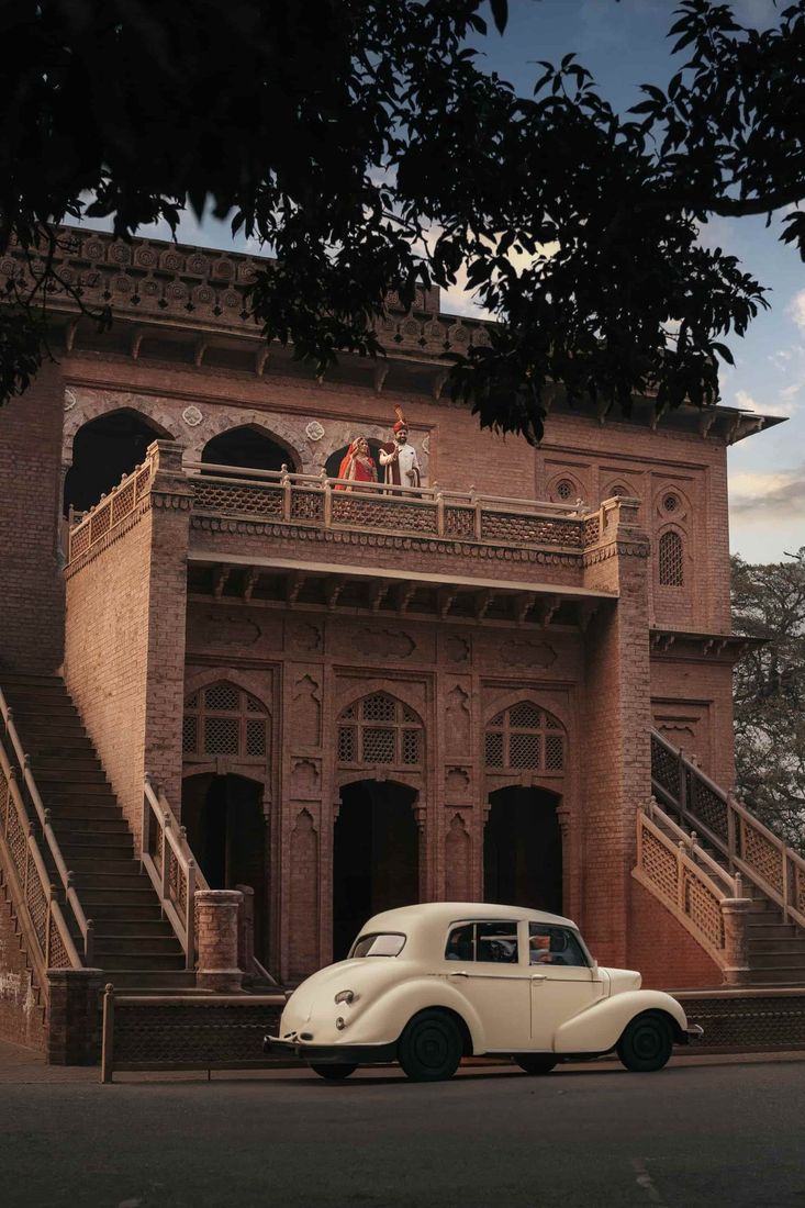 Bride and groom waving from a brick heritage balcony above, a vintage cream classic car parked at street level below