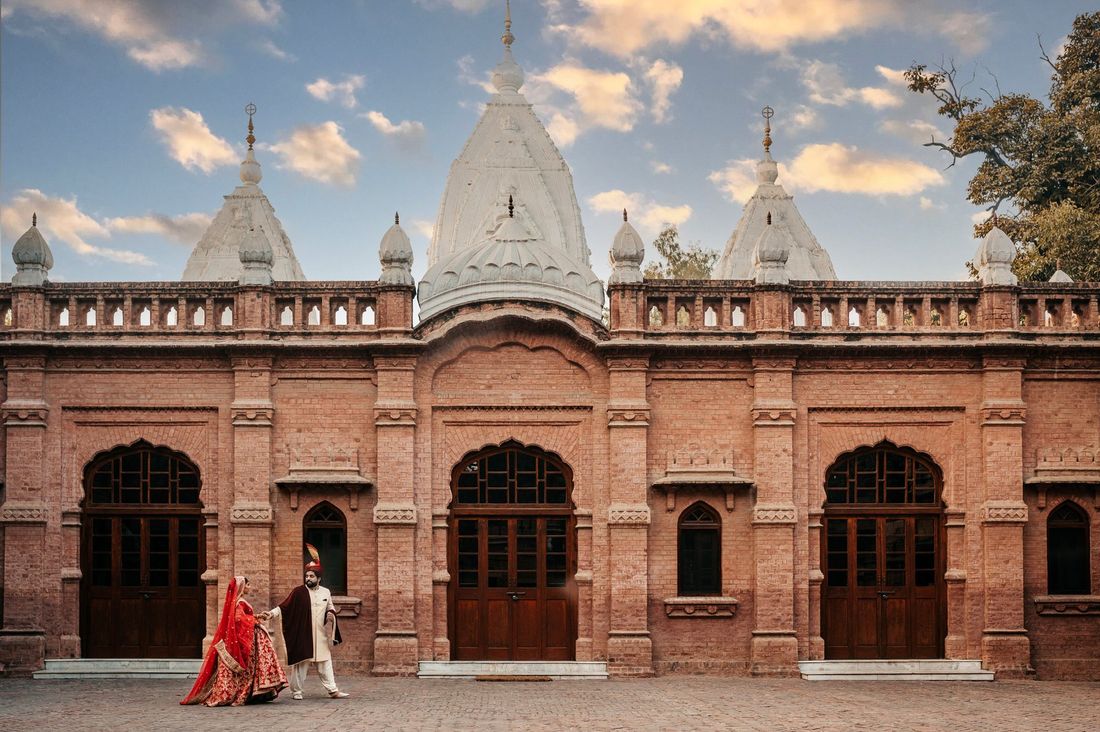 Bride and groom walking hand in hand before a grand brick building with ornate white spires, golden hour sky above