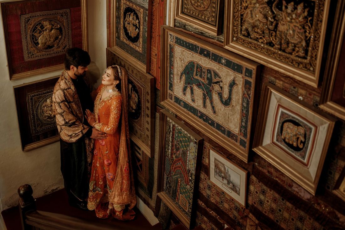 Bride and groom standing close together surrounded by a wall of framed antique tapestries with elephant motifs