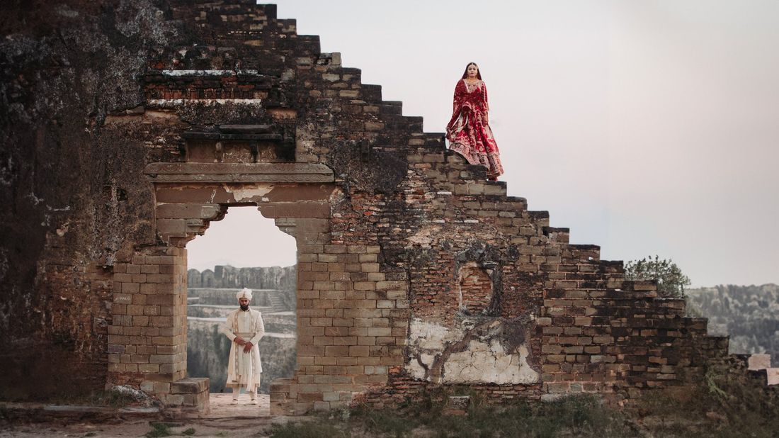 Bride standing atop crumbling stone ruins steps while the groom waits below in a weathered gateway, fort ruins setting