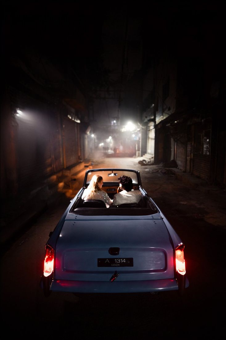 Bride and groom in a vintage convertible driving down a misty, lantern-lit street at night