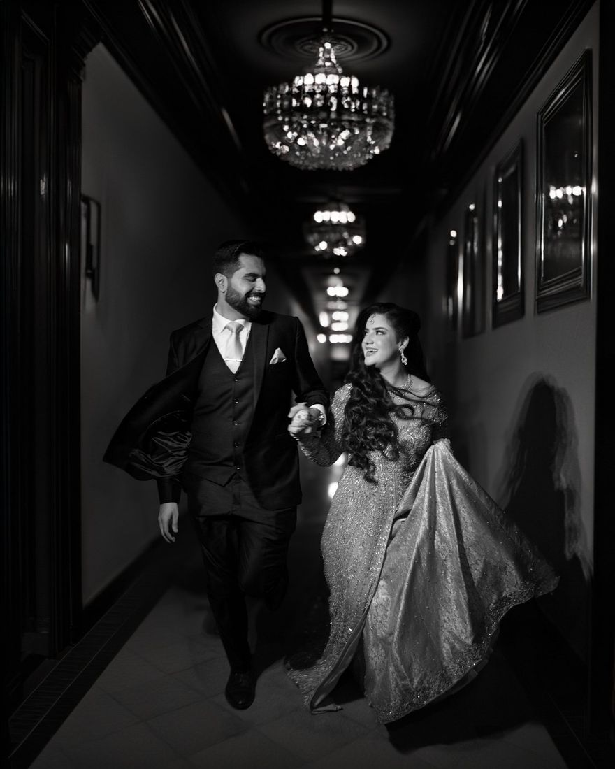 Black-and-white photo of bride and groom laughing as they walk hand in hand down an elegant chandelier-lit hallway