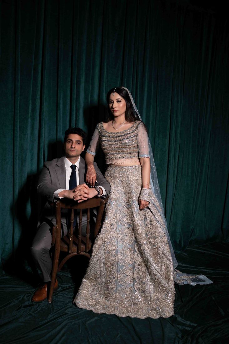 Studio portrait of bride in a powder blue embellished lehenga standing beside the groom seated on a chair, against a dark teal velvet curtain