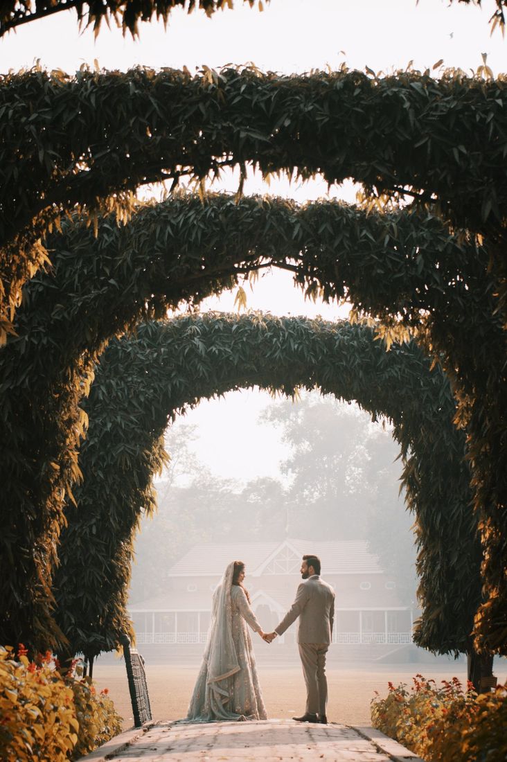 Bride and groom holding hands beneath a series of receding vine-covered archways, a colonial-style building visible in the misty distance