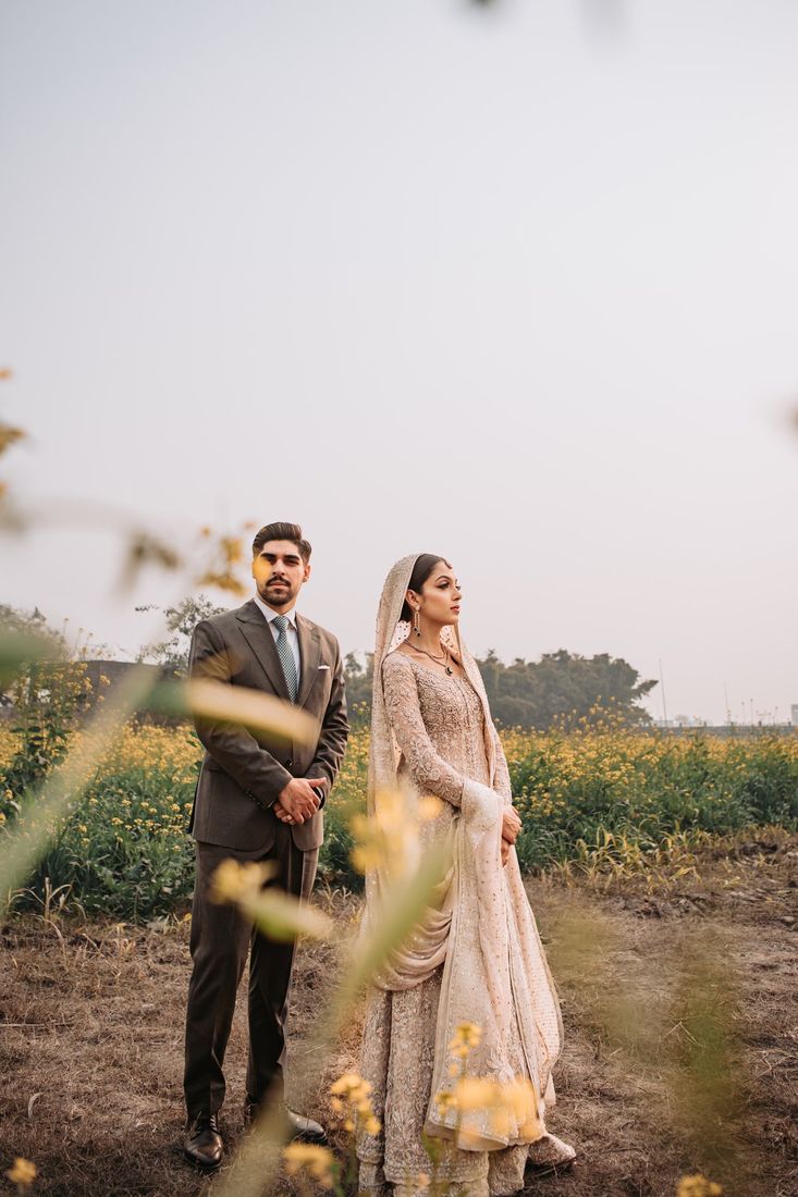 Bride and groom standing together in a mustard flower field, groom in a brown suit and bride in an ivory embellished outfit