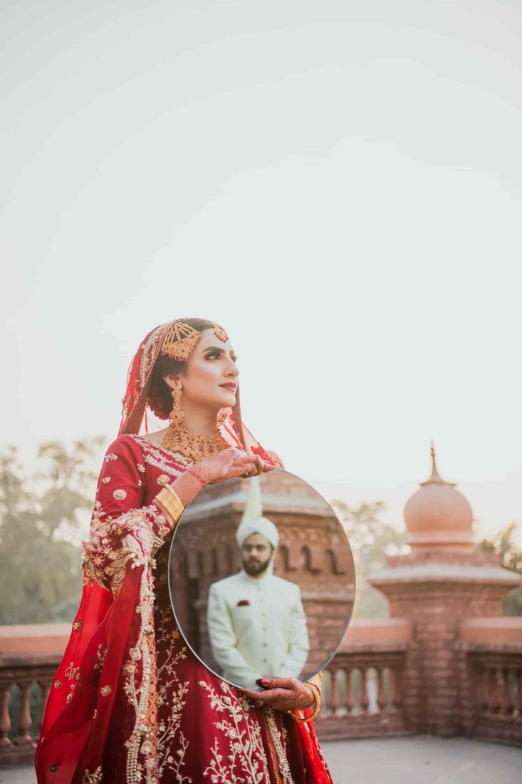 Bride holding a round mirror reflecting the groom in a white sherwani, domed brick architecture behind her