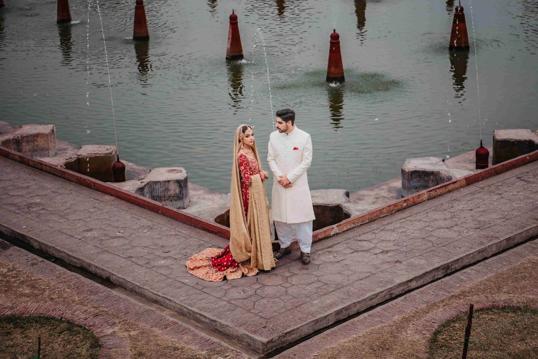 Bride and groom standing beside a still pool with water-jet fountains, bride's gold-bordered red dupatta trailing behind her