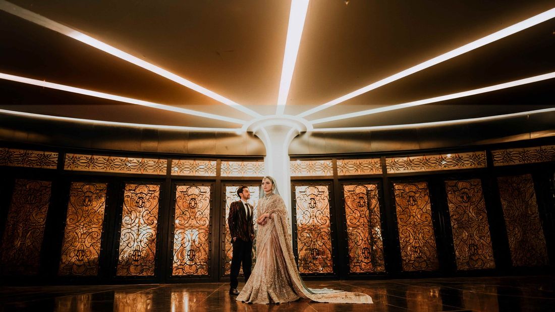 Bride and groom standing beneath a dramatic radial-lit ceiling, ornate gold screens glowing behind them