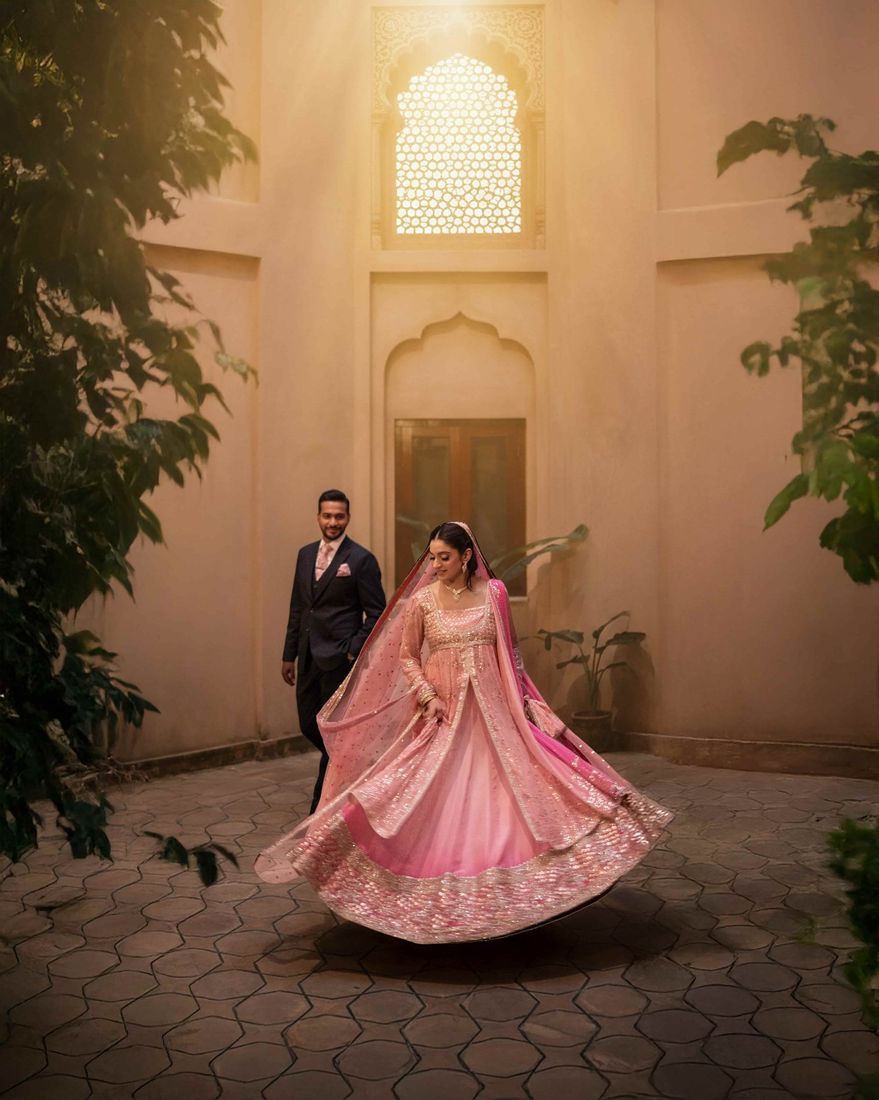 Bride twirling in a pink sequined lehenga as the groom watches, lit by a glowing latticed window behind them