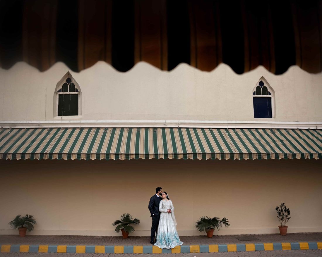 Bride and groom embracing beneath a striped awning and Gothic-arched windows, potted palms lining the wall