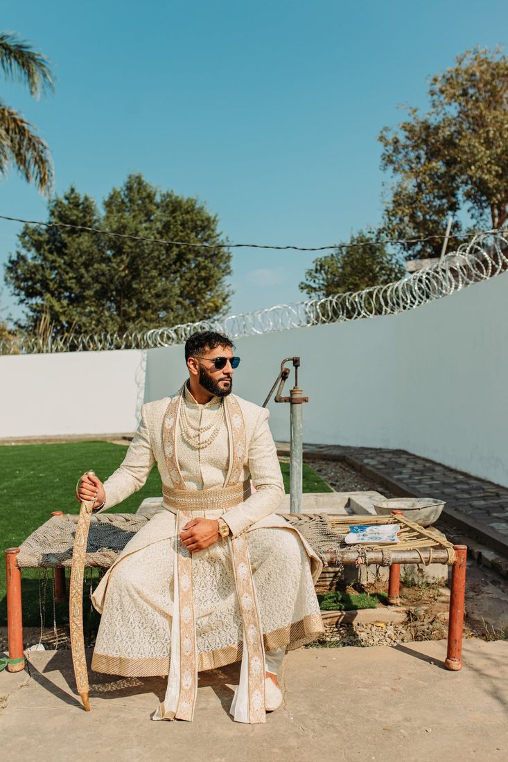 Groom in an ivory sherwani holding a ceremonial sword, seated on a charpai in a candid backyard setting