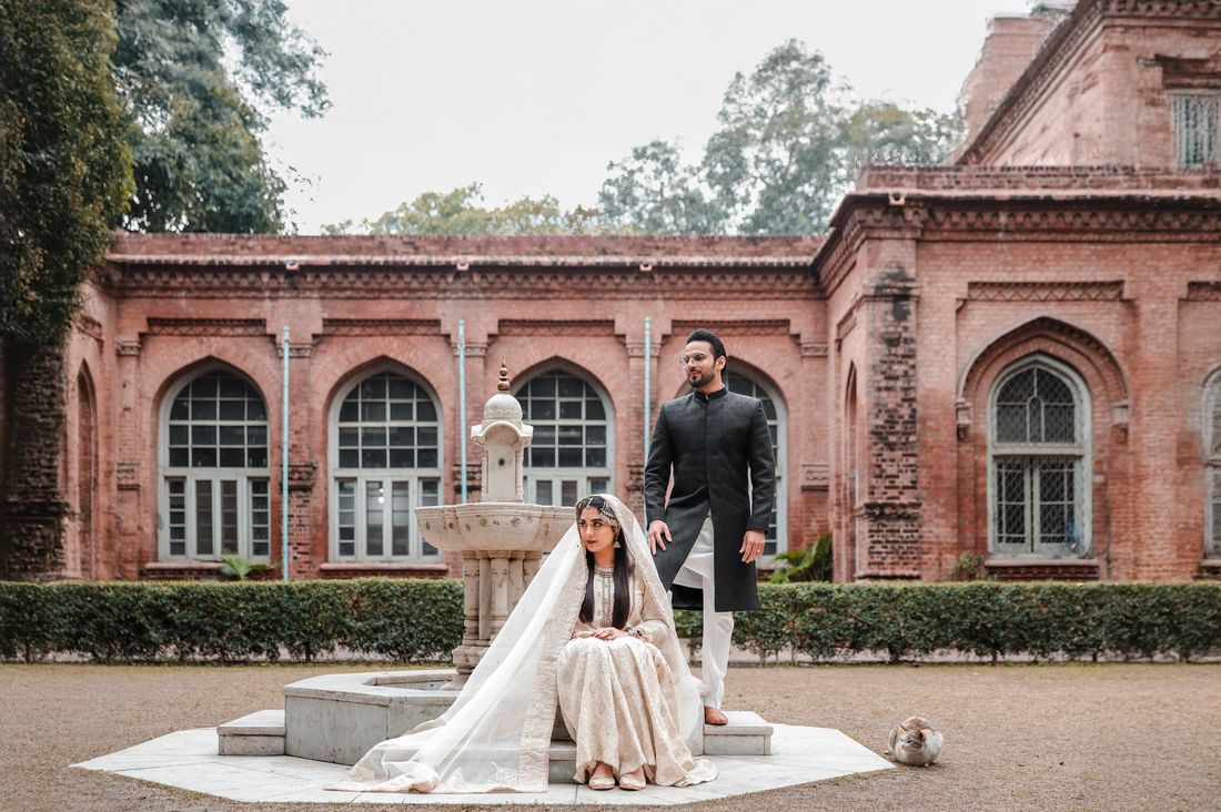 Bride seated beside a marble fountain with the groom standing behind her, in front of a heritage brick building