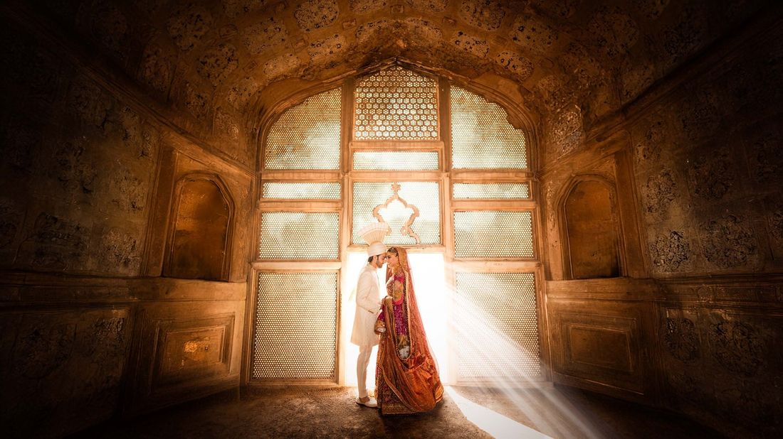 Bride and groom sharing a close moment in a sunlit Mughal archway doorway, golden light streaming in